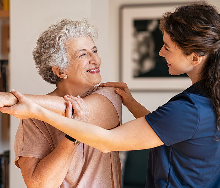 Happy senior woman doing exercise at home with physiotherapist. Old retired lady doing stretching arms at home with the help of a personal trainer during a rehabilitation session.