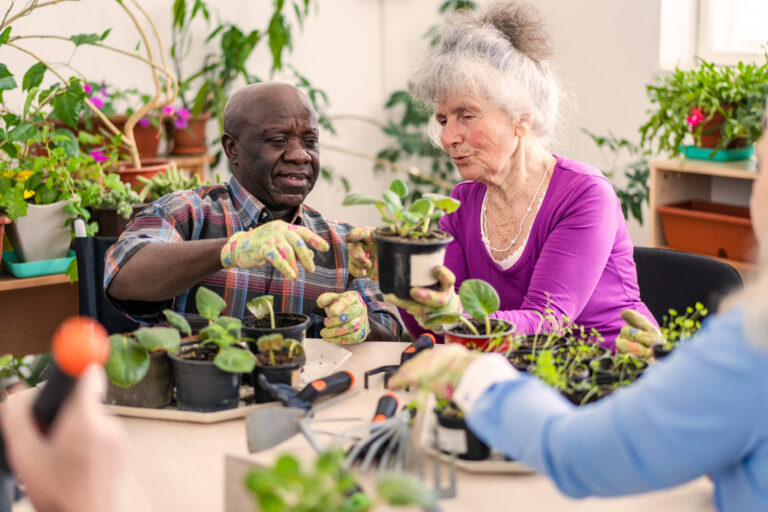 An African-American man wearing gardening gloves is helping an elderly lady sitting next to him with her planting, at a table covered with more pots and plants in a bright sunny room