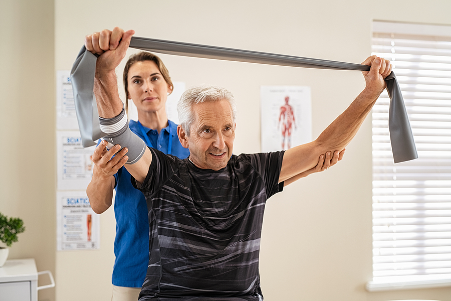 Doctor working with senior patient in rehabilitation center. Trainer assisting senior patient in exercising with resistance band in private clinic.