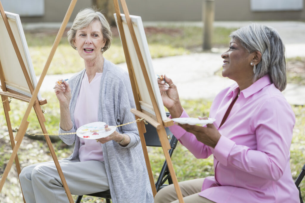 Two multi-ethnic senior women taking an art class outdoors. They are sitting at easels, painting on a canvas. The focus is on the Caucasian woman who is watching her African American friend paint.
