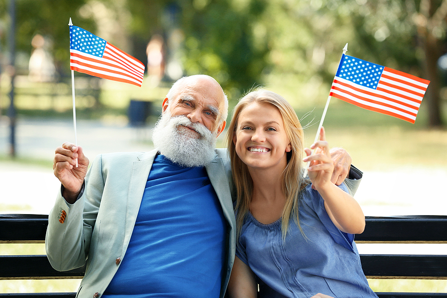 Grandfather and granddaughter sit on a park bench holding American flags and smiling.